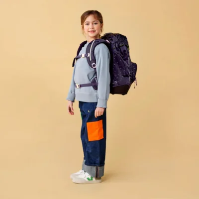 Young girl standing with a large purple backpack, wearing a light blue sweatshirt, dark jeans with an orange patch, and white sneakers.