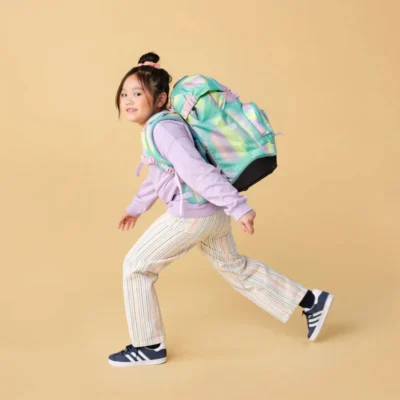 Girl wearing a pastel hoodie and striped pants runs with a colorful backpack against a yellow background.