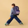 Child wearing a purple starry backpack and denim wide-leg jeans, walking sideways against a beige background.