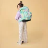 Girl wearing a lavender hoodie and striped pants, carrying a large pastel rainbow backpack against a beige background, looking over her shoulder.