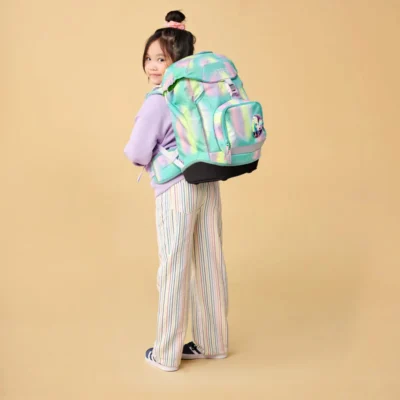 Girl wearing a lavender hoodie and striped pants, carrying a large pastel rainbow backpack against a beige background, looking over her shoulder.